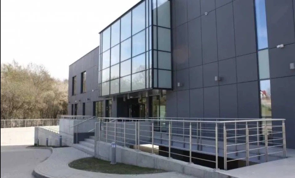 Nowoczesny budynek biurowy z szarą elewacją, przeszklonym wejściem i podjazdem dla niepełnosprawnych wyłożonym kostką brukową. Widoczne drzewa w tle.