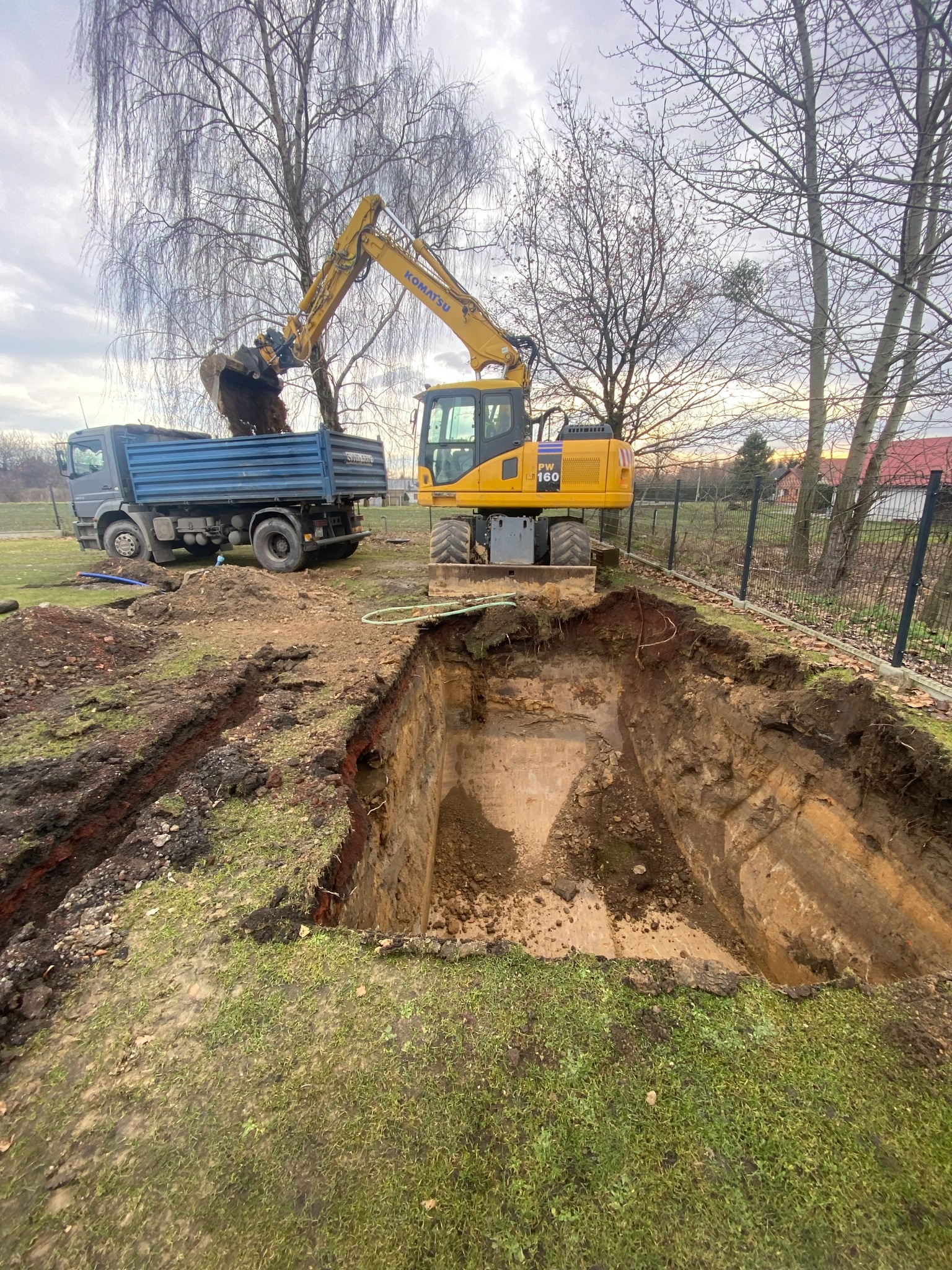 Żółta koparka Komatsu PW160 ładuje ziemię do niebieskiej ciężarówki. Wykopany dół w ziemi, trawa, drzewa bez liści i ogrodzenie w tle.