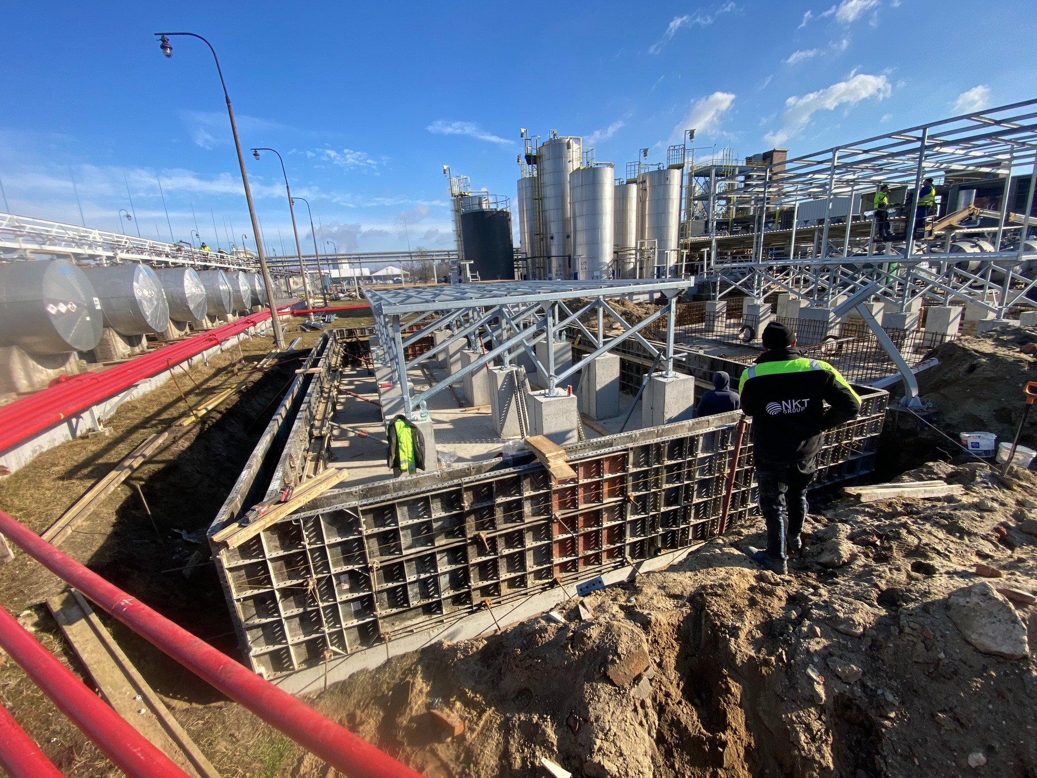 Wykop z szalunkiem i zbrojeniem fundamentów pod konstrukcję przemysłową. W tle zbiorniki i stalowa konstrukcja. Prace ziemne i fundamentowe w toku.