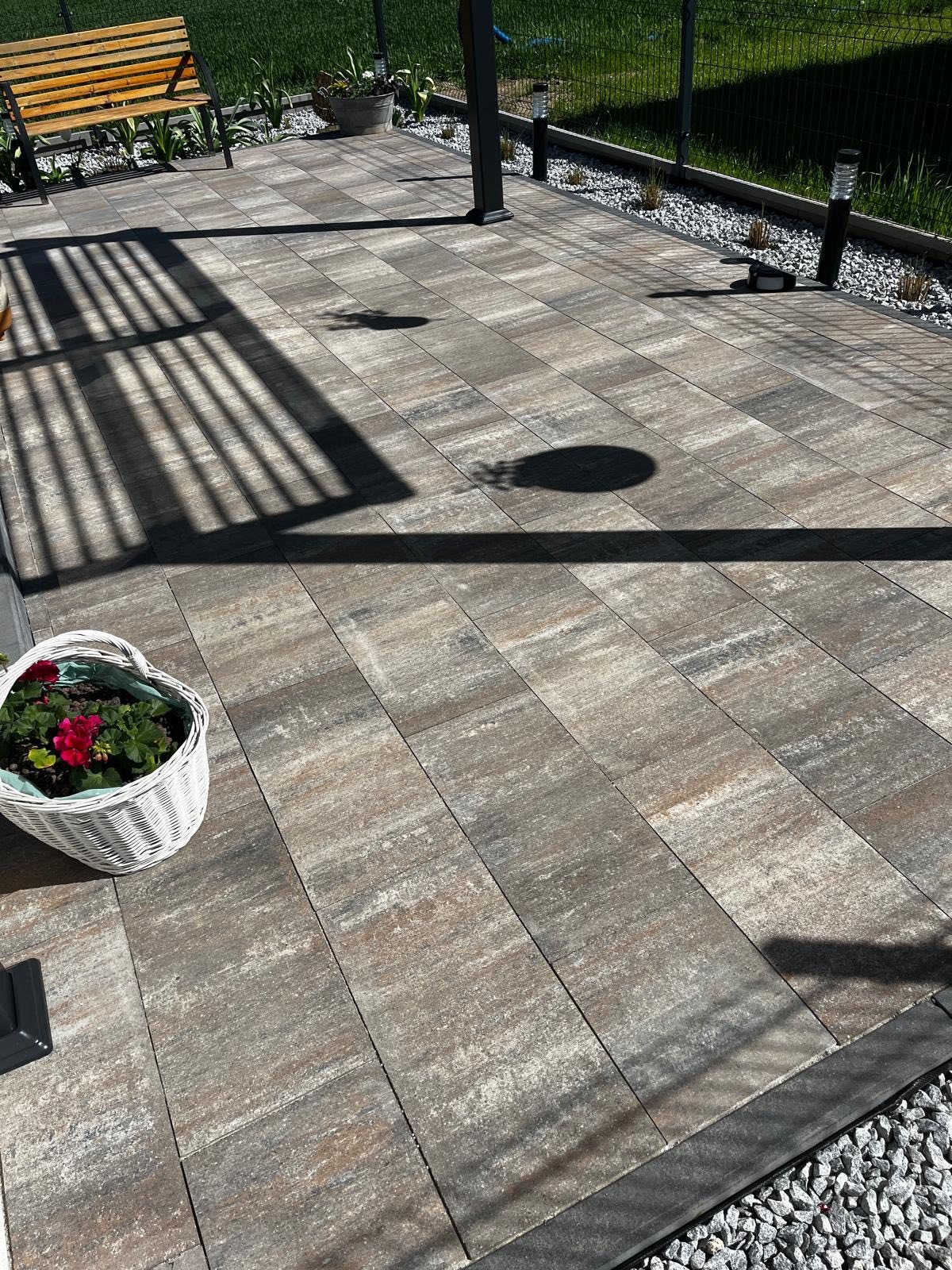 Ułożona kostka brukowa na posesji w Jastrzębiu-Zdroju. Widoczny wzór ułożenia, rabata z kamienia, ławka i cień pergoli. Koszyk z kwiatami w rogu kadru.