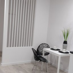 Grzegorz Jaszczuk - Jasne wnętrze: panelowa ścianka działowa, podłoga imitująca drewno, stół z dekoracjami i krzesła o ażurowych oparciach. Minimalistyczny design.