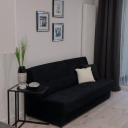 Grzegorz Jaszczuk - Nowoczesny salon: czarna sofa z poduszkami, szklany stolik z dekoracjami, drewniana podłoga, ściana z trzema obrazami w srebrnych ramkach i zasłony.