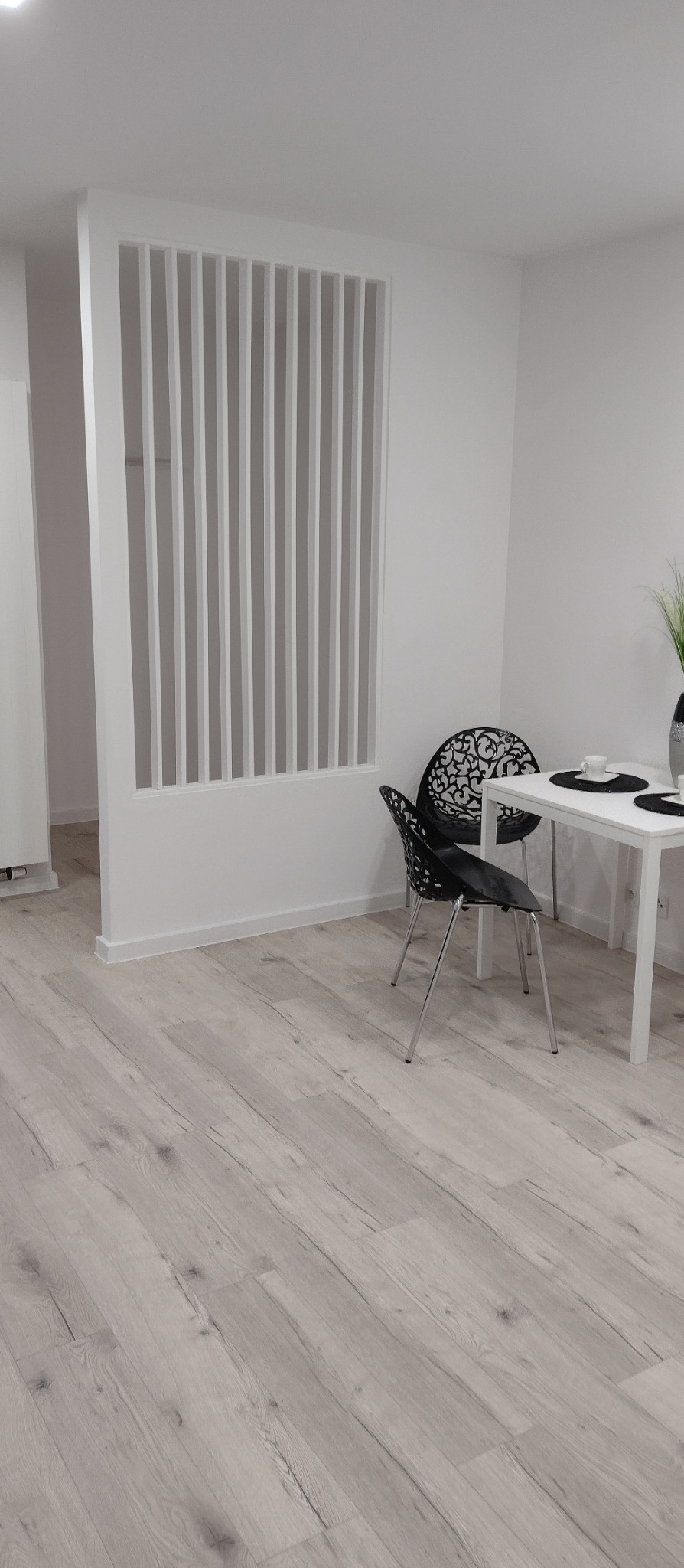 Białe lamele działowe w nowoczesnym wnętrzu z podłogą imitującą jasne drewno, czarne krzesła ażurowe i stół. Minimalistyczny design, jasne kolory, naturalne światło.