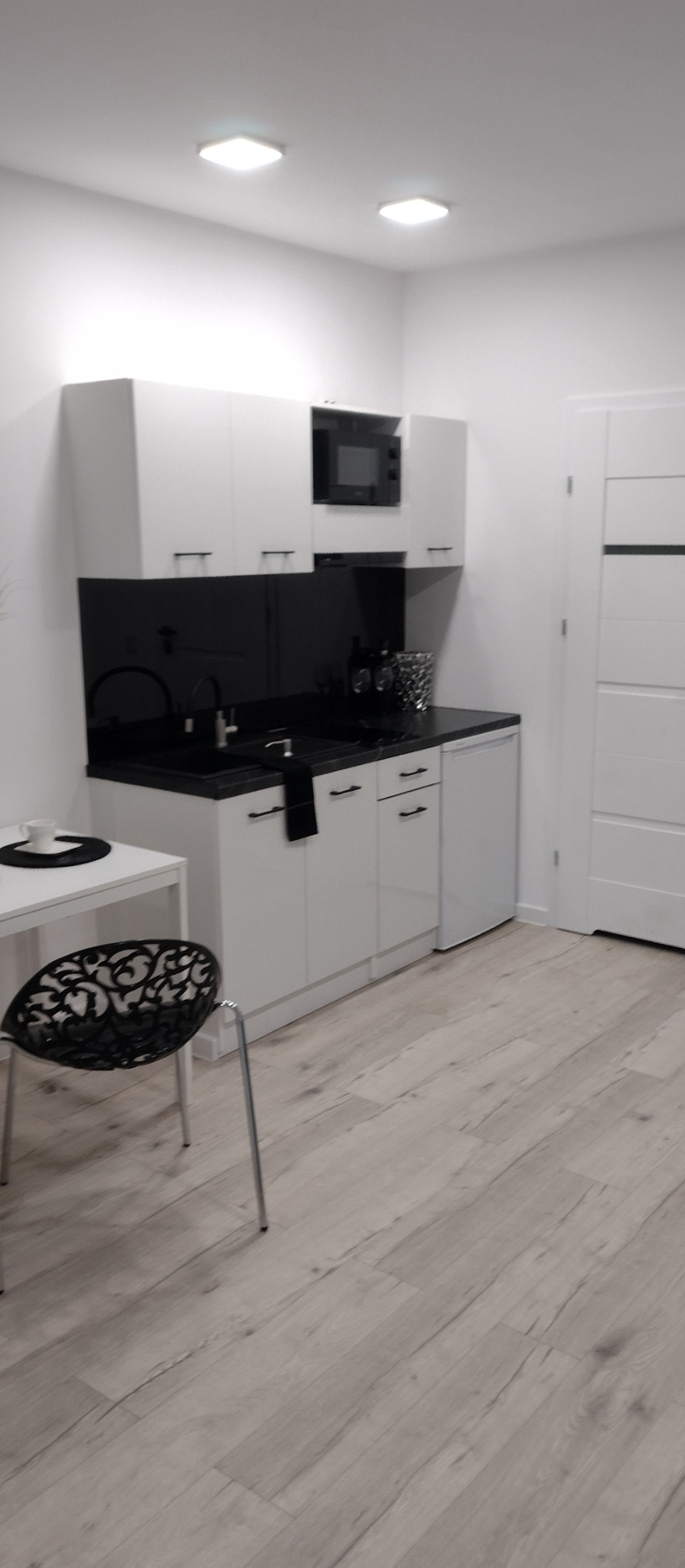 Nowoczesna, minimalistyczna kuchnia w bieli i czerni. Widoczne białe szafki, czarny blat, dekoracyjny krzesło i panele podłogowe. Całość utrzymana w jasnej tonacji.
