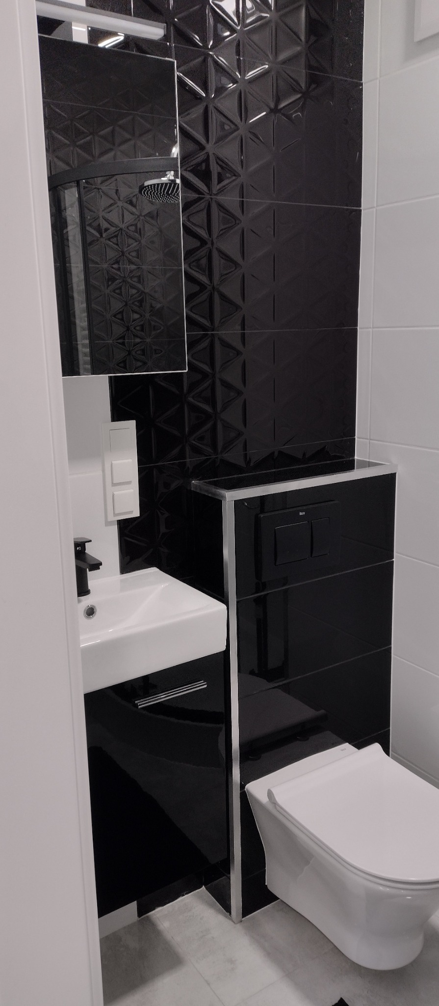 Nowoczesna łazienka: czarne, trójwymiarowe płytki, biała umywalka z szafką i toaleta. W lustrze odbija się deszczownica. Całość w minimalistycznym stylu.