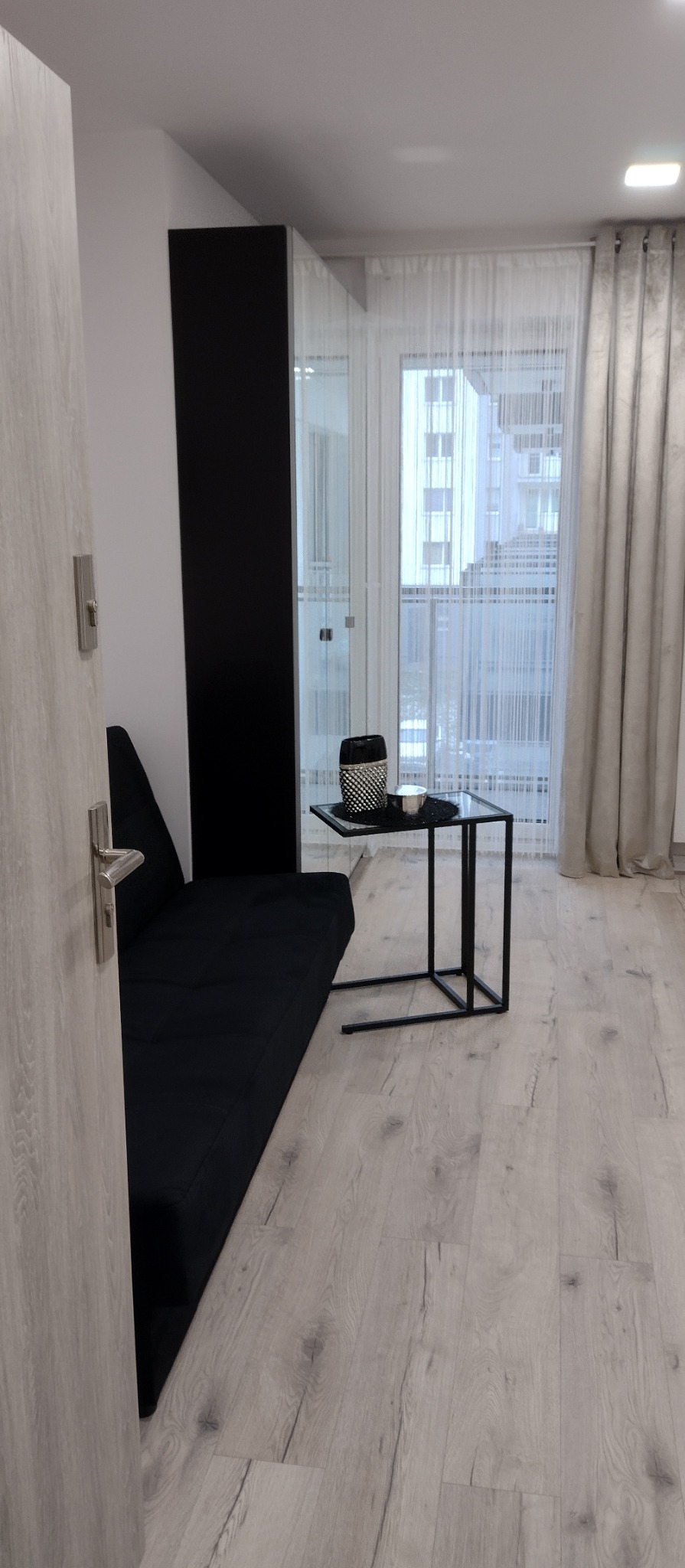 Nowoczesny salon w Rosanowie: czarna sofa, szklany stolik i jasna podłoga. Minimalistyczny design z widokiem na okno i firany.