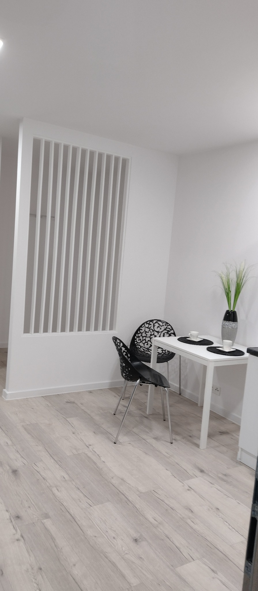 Jasne wnętrze: panelowa ścianka działowa, podłoga imitująca drewno, stół z dekoracjami i krzesła o ażurowych oparciach. Minimalistyczny design.