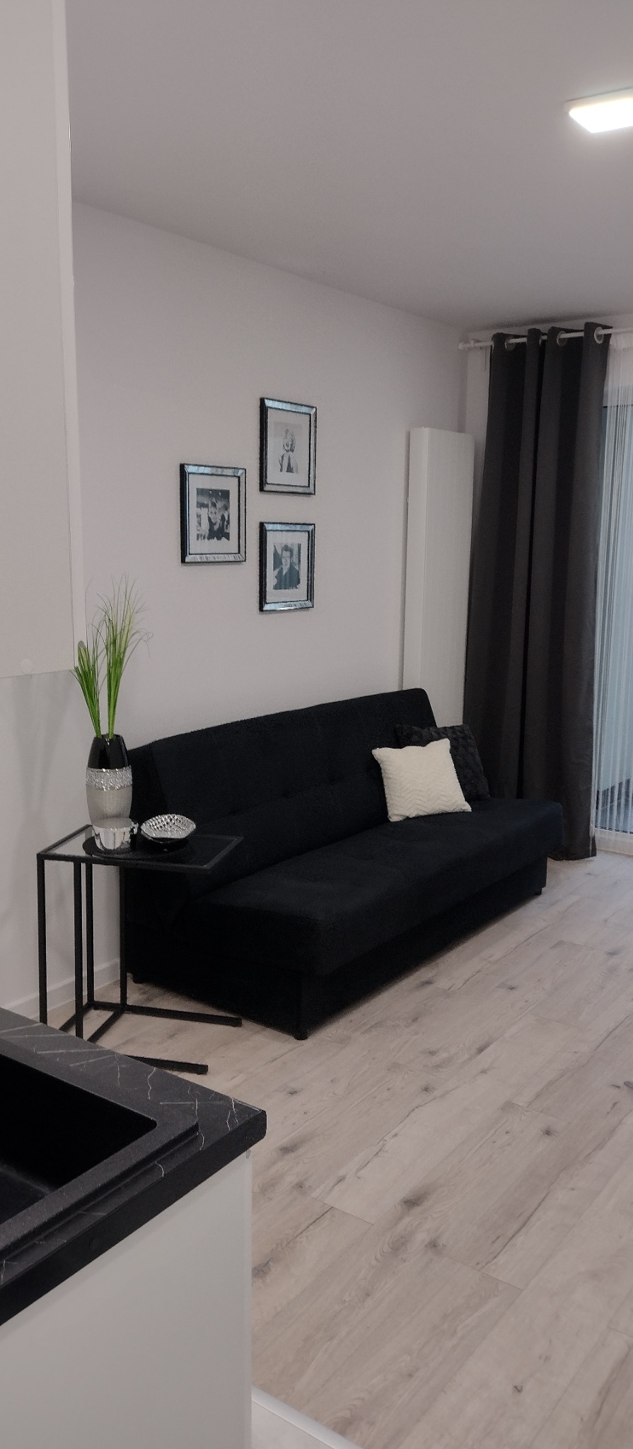 Nowoczesny salon: czarna sofa z poduszkami, szklany stolik z dekoracjami, drewniana podłoga, ściana z trzema obrazami w srebrnych ramkach i zasłony.