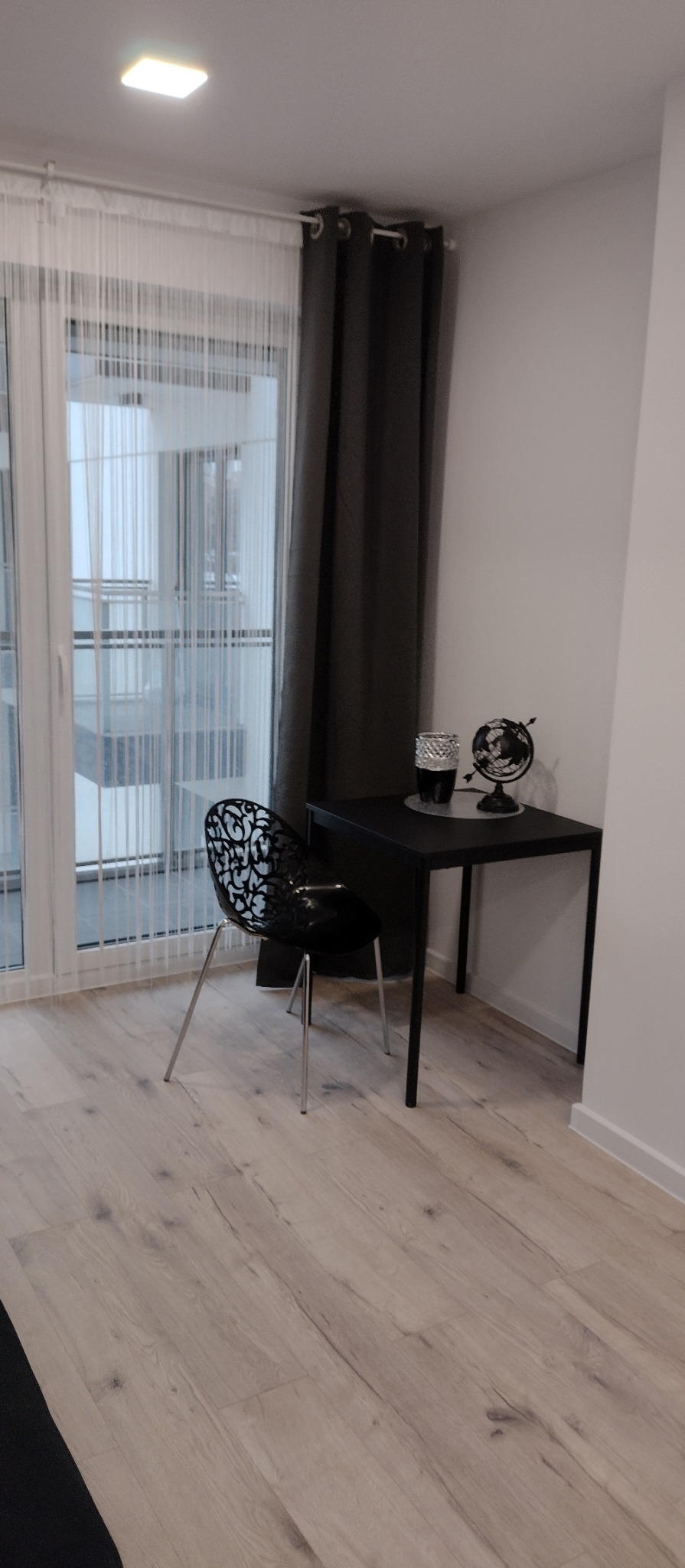 Jasne panele podłogowe, czarne krzesło z ażurowym oparciem i biurko z globusem w minimalistycznym, nowocześnie urządzonym pokoju z balkonem.