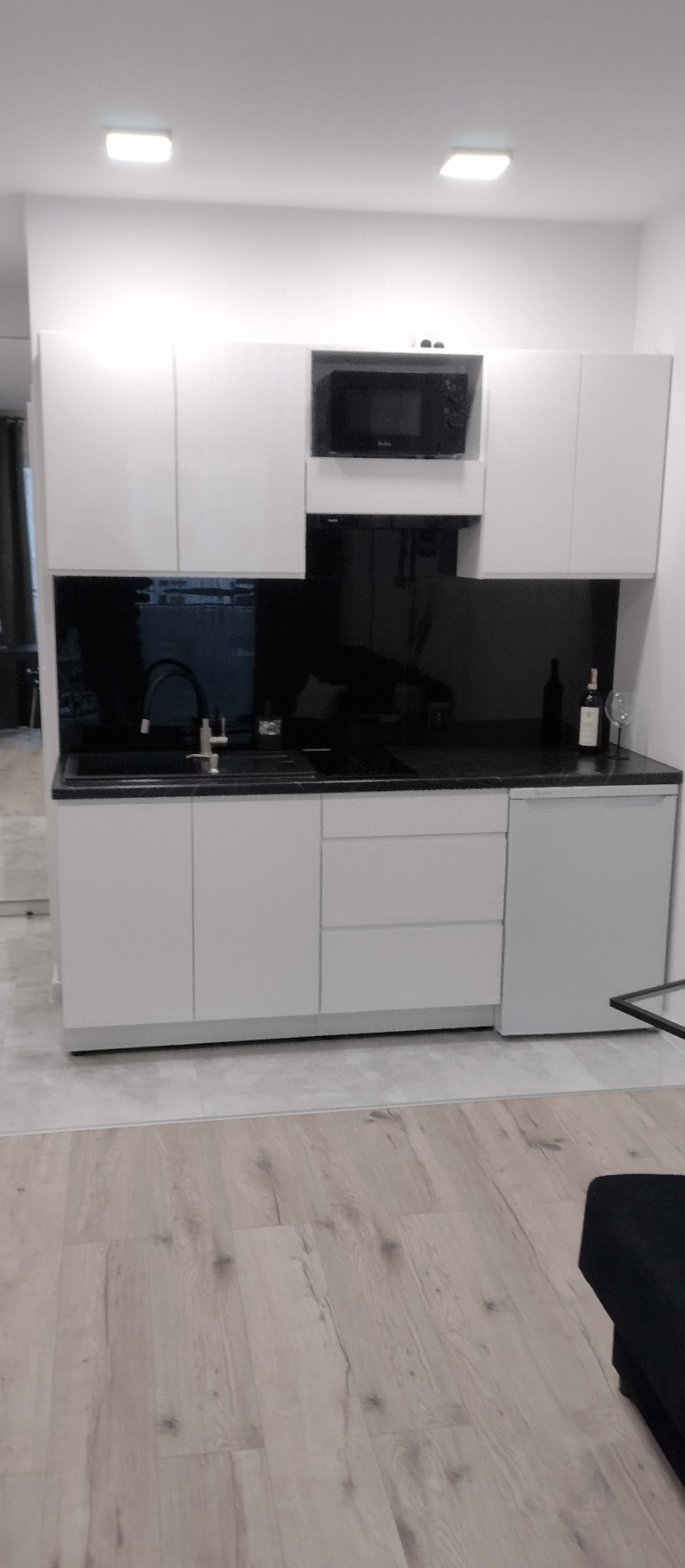 Nowoczesna, minimalistyczna kuchnia z białymi szafkami i czarnym blatem. Widoczna butelka wina i kieliszek. Podłoga wyłożona panelami imitującymi drewno.