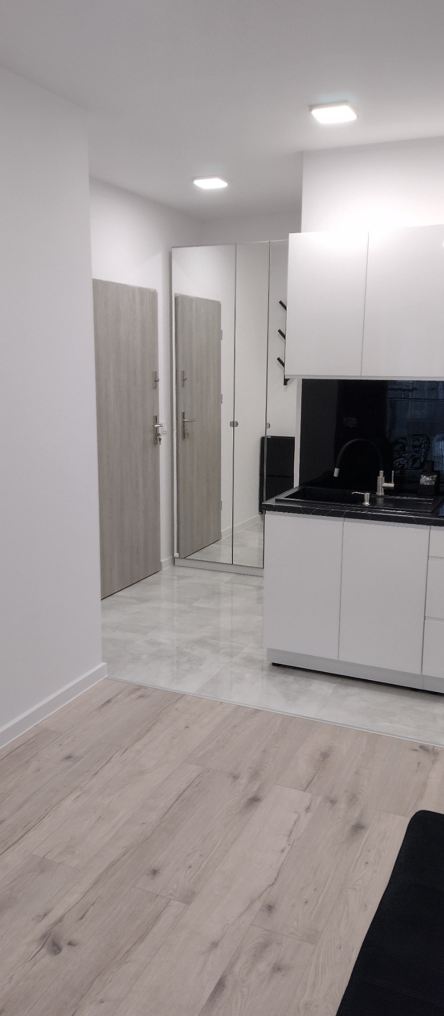 Nowoczesne wykończenie wnętrza: biała zabudowa kuchenna z czarnym blatem, drzwi z szarego drewna i podłoga z płytek imitujących drewno w jasnej tonacji.