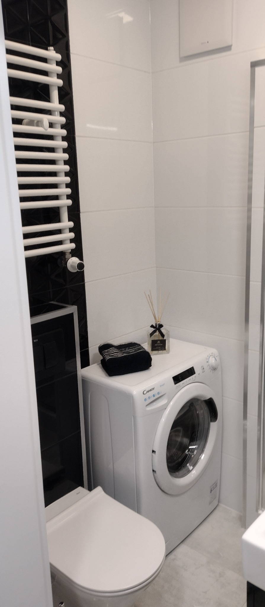 Nowoczesna, wykończona łazienka w bieli i czerni z pralką Candy, grzejnikiem drabinkowym i czarnymi płytkami dekoracyjnymi na ścianie. Minimalistyczny design.