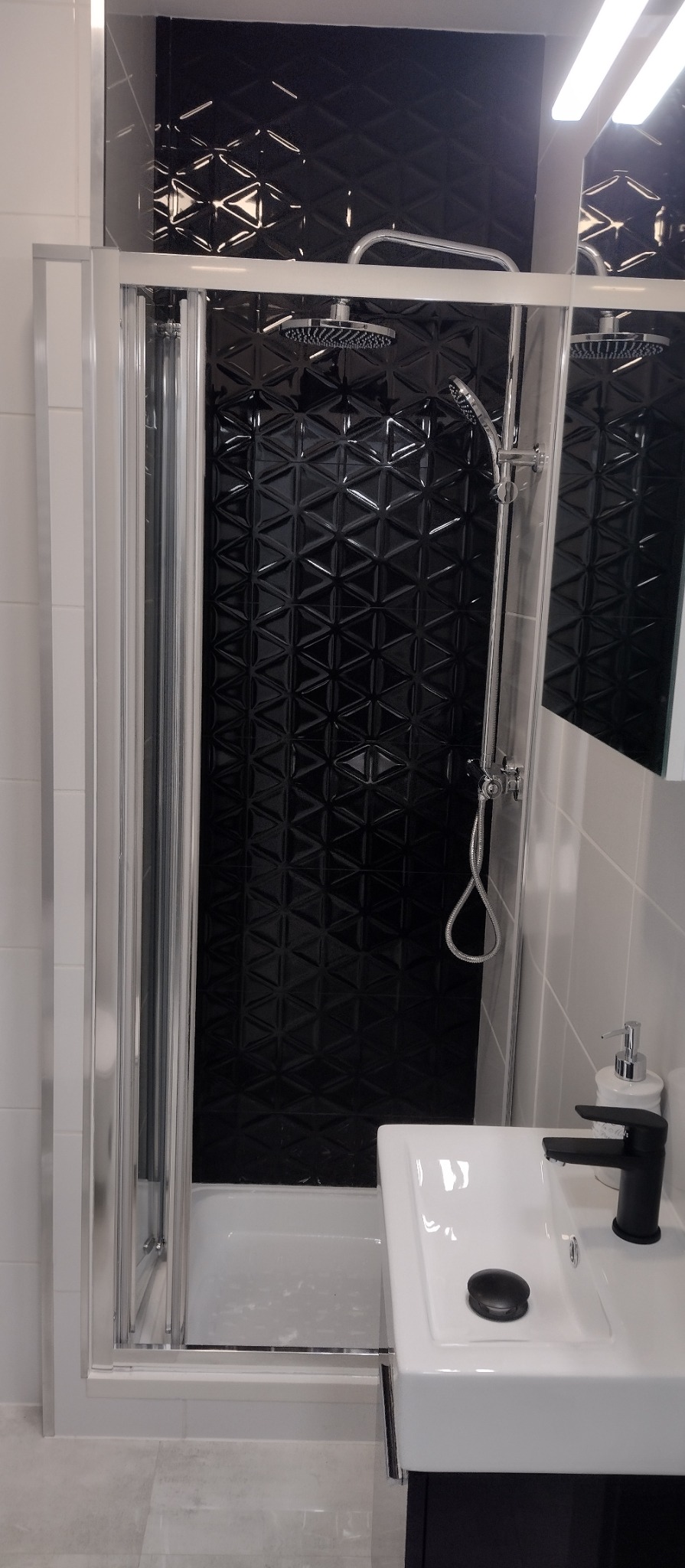 Nowoczesna kabina prysznicowa z czarną, trójwymiarową glazurą i białą armaturą. Umywalka z czarną baterią i dozownik mydła. Minimalistyczny design łazienki.