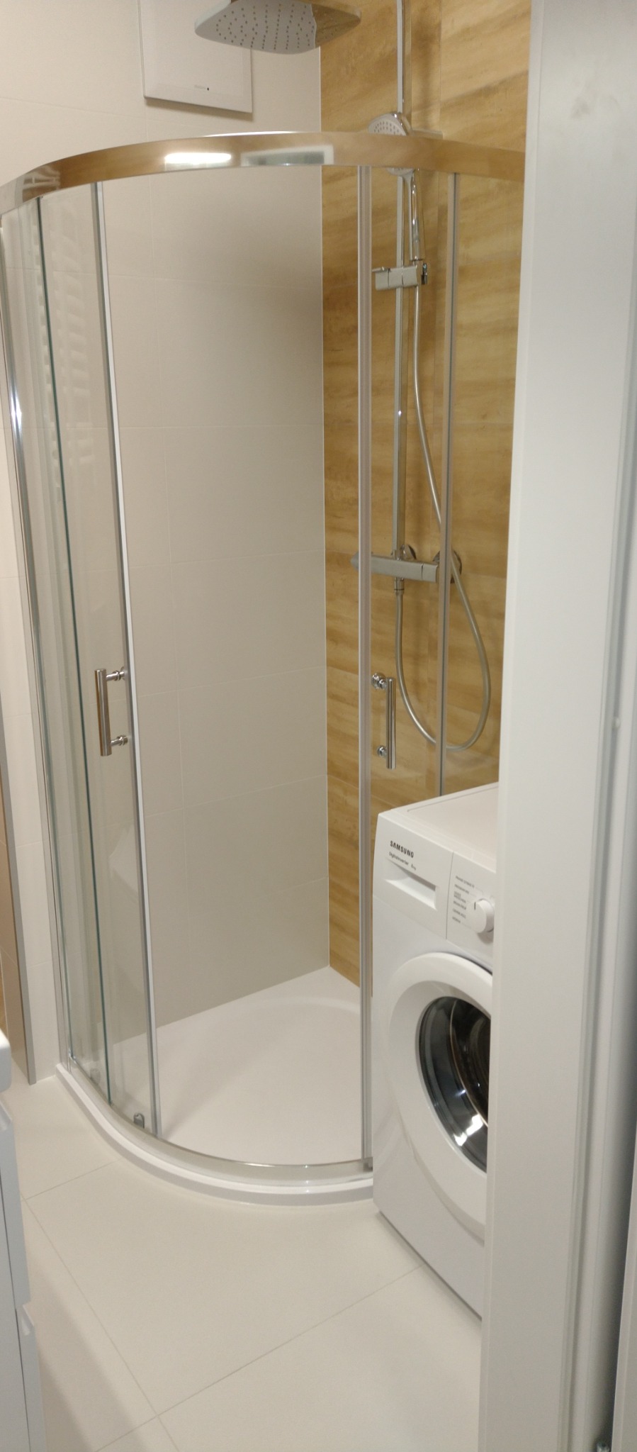 Narożna kabina prysznicowa z chromowanymi profilami i deszczownicą, obok pralka Samsung. Beżowe płytki na ścianach. Jasna, nowoczesna łazienka po remoncie.