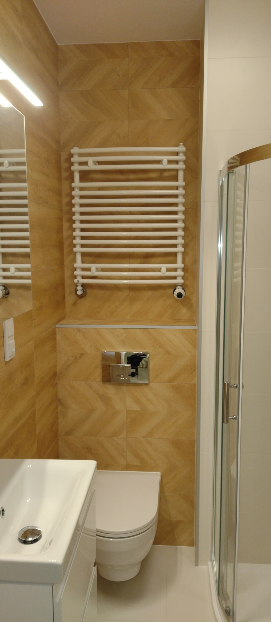 Beżowa łazienka po remoncie: grzejnik drabinkowy, toaleta podwieszana, kabina prysznicowa i umywalka. Ułożone płytki z wzorem jodełki. Nowoczesny design.