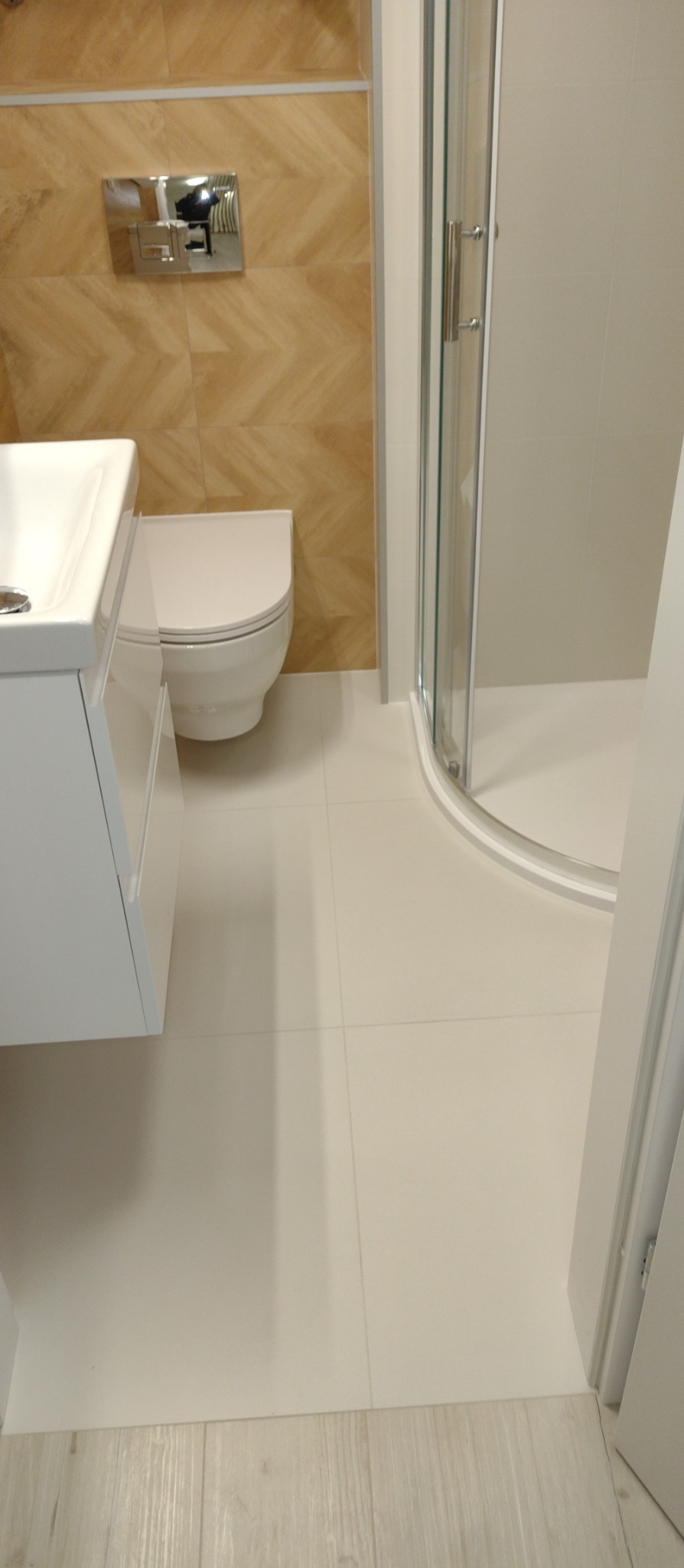 Wykończona łazienka z kabiną prysznicową, toaletą i umywalką. Na ścianie płytki w jodełkę. Jasne płytki na podłodze. Nowoczesny design.