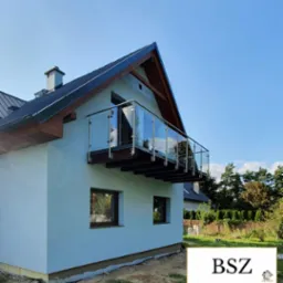 Nowoczesny balkon z przeszkloną balustradą w domu jednorodzinnym, widok z dołu na tle błękitnego nieba. Elementy drewniane wykończenia dachu. Minimalistyczny design.