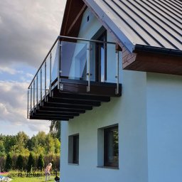 ZAB-DEKOR Arkadiusz Zabdyr - Nowoczesny balkon z szklaną balustradą i drewnianym podestem, zamontowany przy jasnym budynku z ciemnym dachem. W tle zieleń ogrodu i bawiąca się dziewczynka.