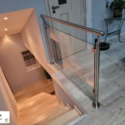 ZAB-DEKOR Arkadiusz Zabdyr - Szklana balustrada ze stalową poręczą na schodach z jasnego drewna, widok z góry. W tle drzwi i stolik z rośliną doniczkową.