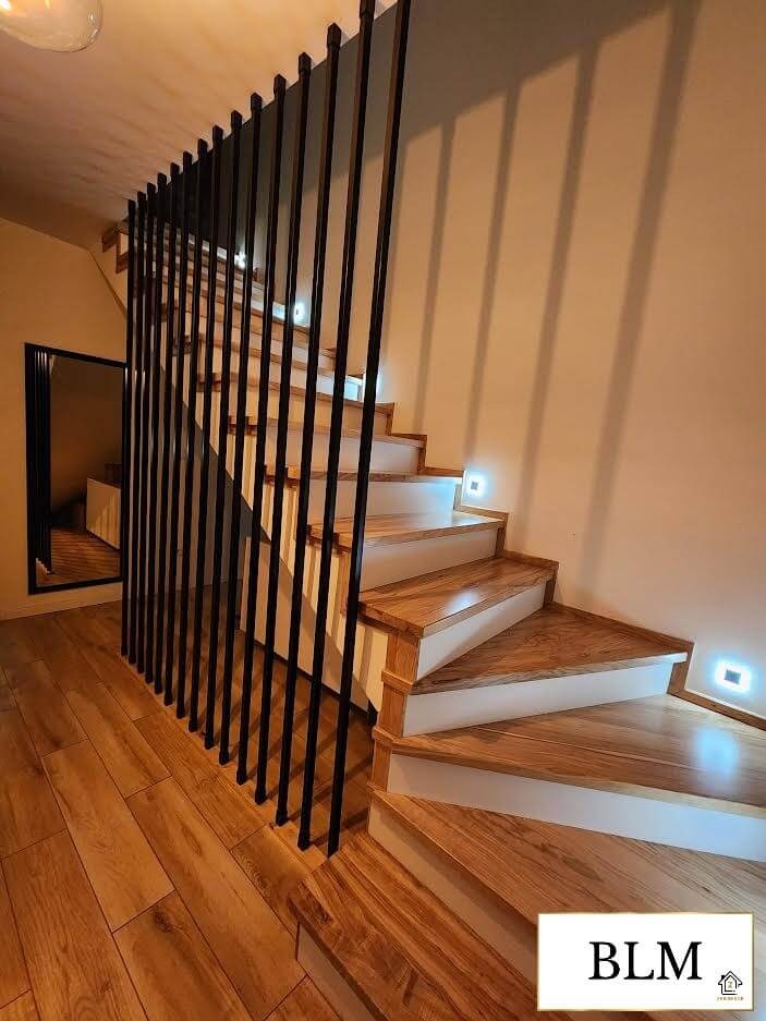 Drewniane schody z podświetleniem LED na stopniach, zabezpieczone czarną, metalową konstrukcją. Lustro w tle. Parkiet na podłodze.