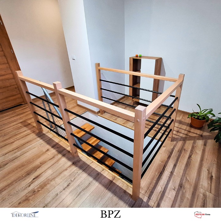 Klatka schodowa z jasną drewnianą balustradą i czarnymi, metalowymi prętami, widziana z góry. W tle drewniany regał i roślina doniczkowa.