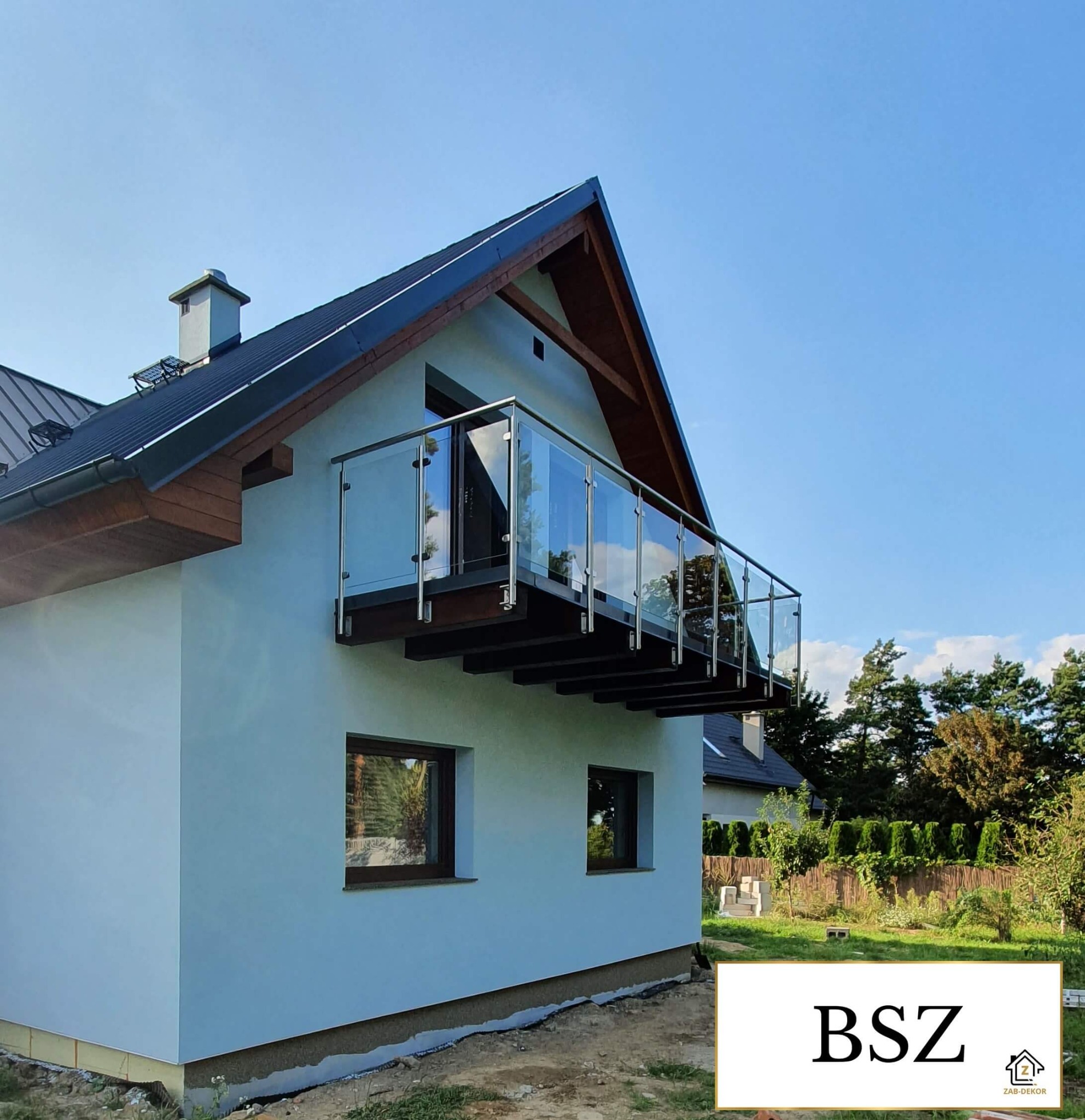 Nowoczesny balkon z przeszkloną balustradą w domu jednorodzinnym, widok z dołu na tle błękitnego nieba. Elementy drewniane wykończenia dachu. Minimalistyczny design.