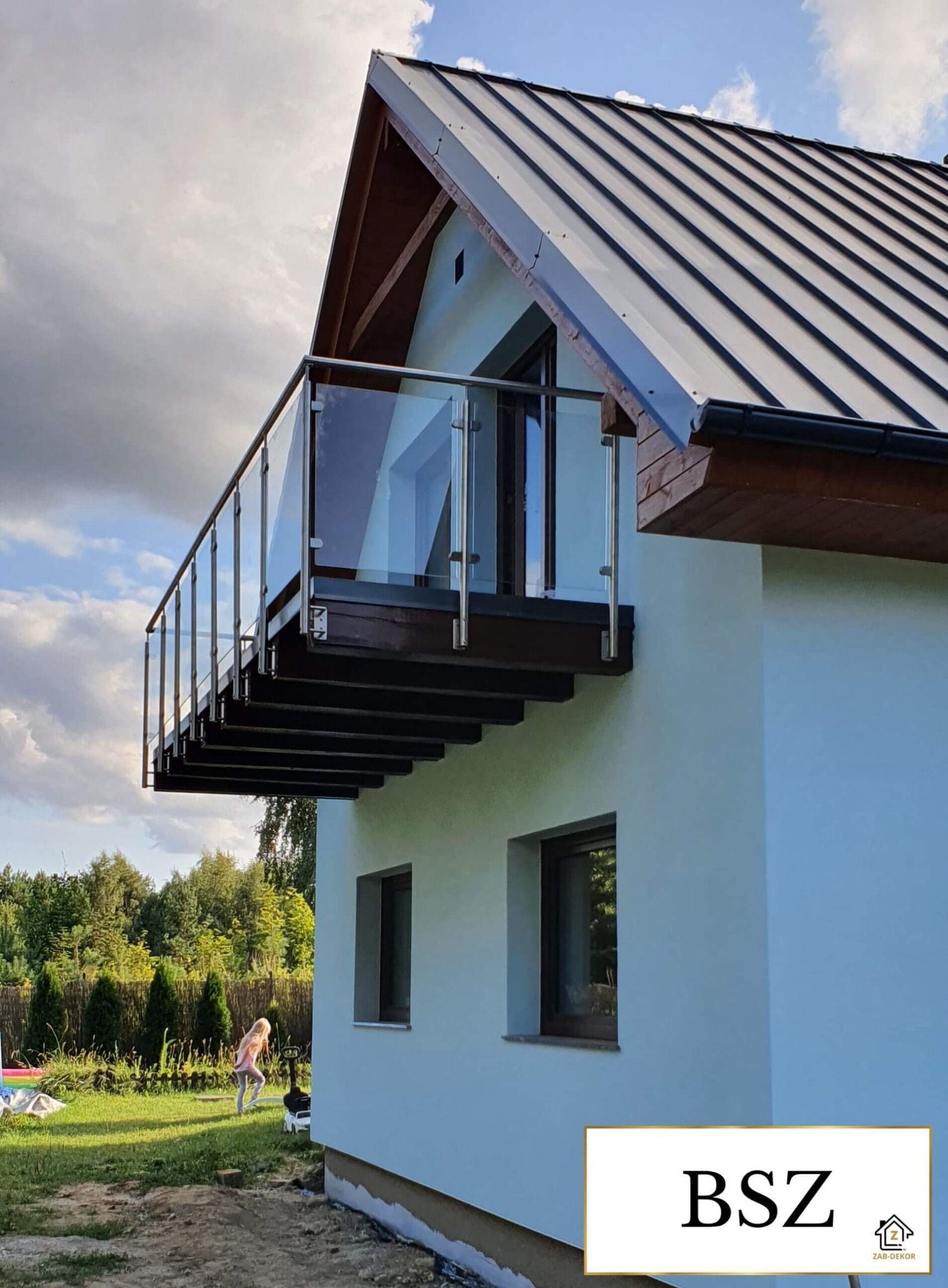 Nowoczesny balkon z szklaną balustradą i drewnianym podestem, zamontowany przy jasnym budynku z ciemnym dachem. W tle zieleń ogrodu i bawiąca się dziewczynka.
