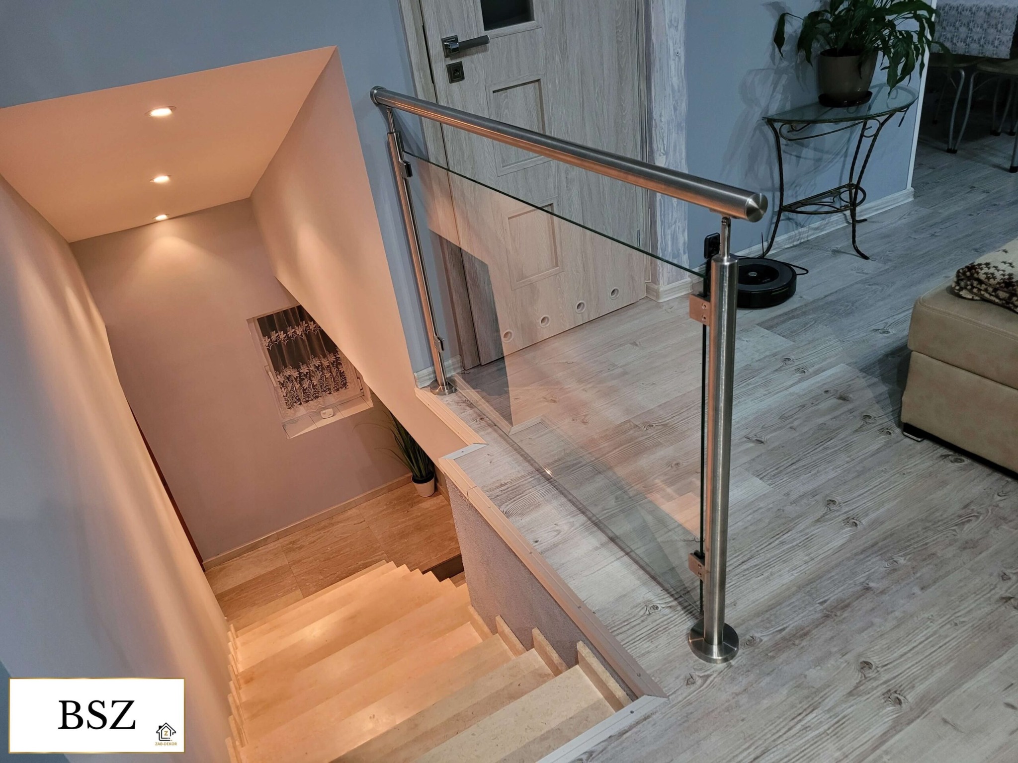 Szklana balustrada ze stalową poręczą na schodach z jasnego drewna, widok z góry. W tle drzwi i stolik z rośliną doniczkową.