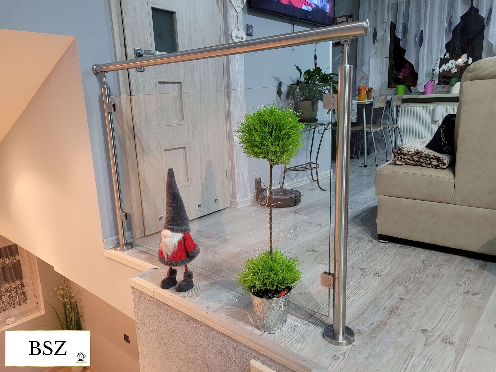 Nowoczesna balustrada szklana ze stali nierdzewnej, zabezpieczająca schody. W tle widoczny wystrój wnętrza domu z dekoracjami i meblami.