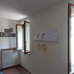 Klimatyzacja Monoblok - Przygotowanie do montażu klimatyzacji: szablon na ścianie z zaznaczonymi otworami, widoczne nawierty. W tle okno z zasłoną i grzejnik.