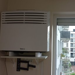 Klimatyzacja Monoblok - Biały klimatyzator UNICO zamontowany na ścianie w pokoju, widok z okna na budynki mieszkalne. Minimalistyczny design, jasne wnętrze.