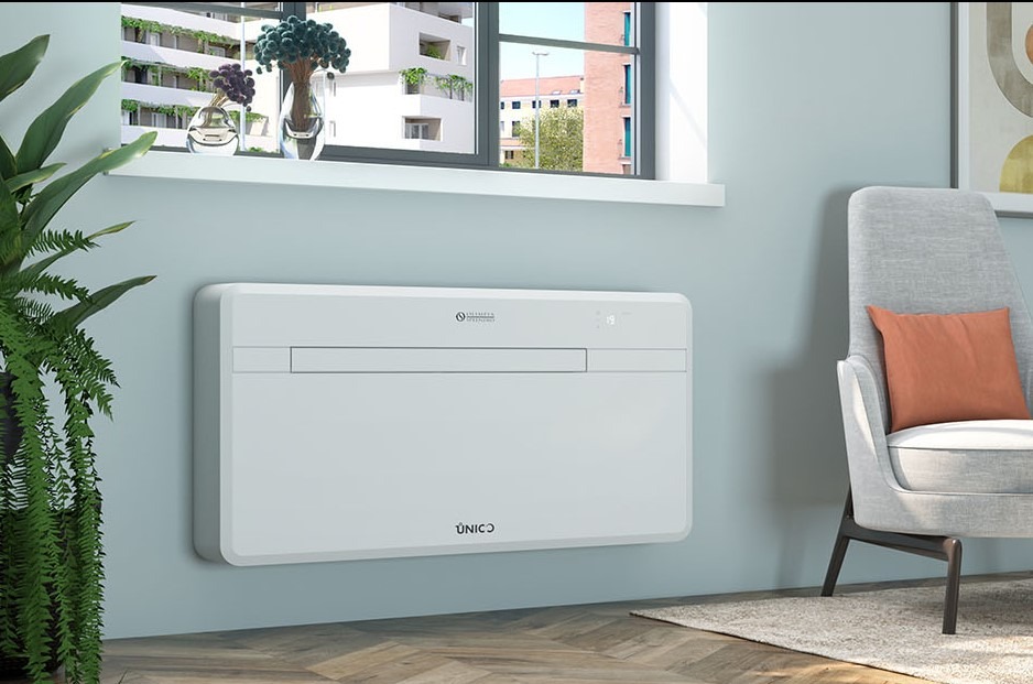 Biały, ścienny klimatyzator Unico w nowoczesnym wnętrzu. Widok na okno z roślinami i budynki w tle. Minimalistyczny design, szary fotel z poduszką.