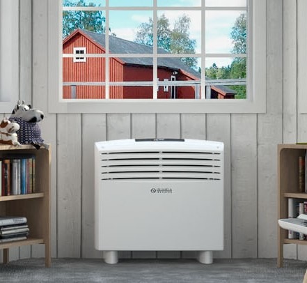 Biały przenośny klimatyzator NORDMANN stojący w pokoju z drewnianą boazerią, książkami na półkach i widokiem na czerwony dom za oknem.