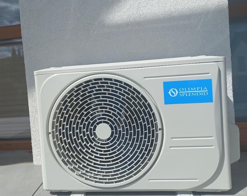 Biała jednostka zewnętrzna klimatyzacji Olimpia Splendid zamontowana na ścianie budynku, widoczna z bliska. Szara podstawa, błękitne logo.