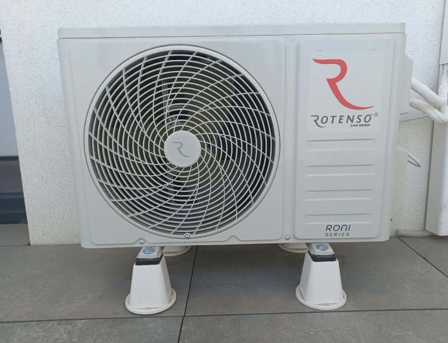 Biały klimatyzator Rotenso Roni Series, umieszczony na specjalnych podkładkach, z widocznym logo i wentylatorem, na tle ściany i szarej podłogi.