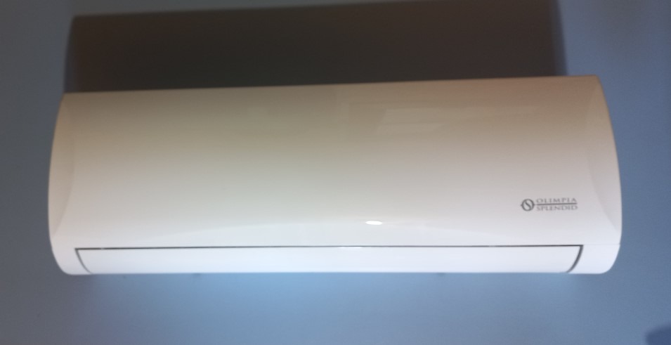 Biały klimatyzator Olimpia Splendid zamontowany na niebieskiej ścianie, oświetlony od dołu, widoczny z przodu, w tle cień na suficie.