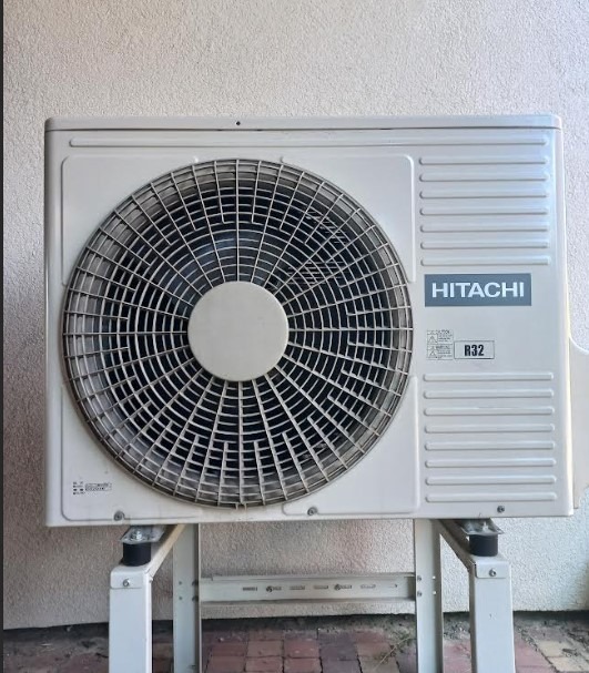 Biała, zewnętrzna jednostka klimatyzacji Hitachi R32, zamontowana na metalowym stojaku, na tle jasnej ściany i fragmentu czerwonej kostki brukowej.