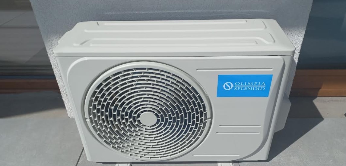 Kompaktowy klimatyzator Olimpica Splendid, zamontowany na zewnątrz budynku, z widoczną obudową i niebieskim logo, na tle jasnej ściany i szarej podłogi.