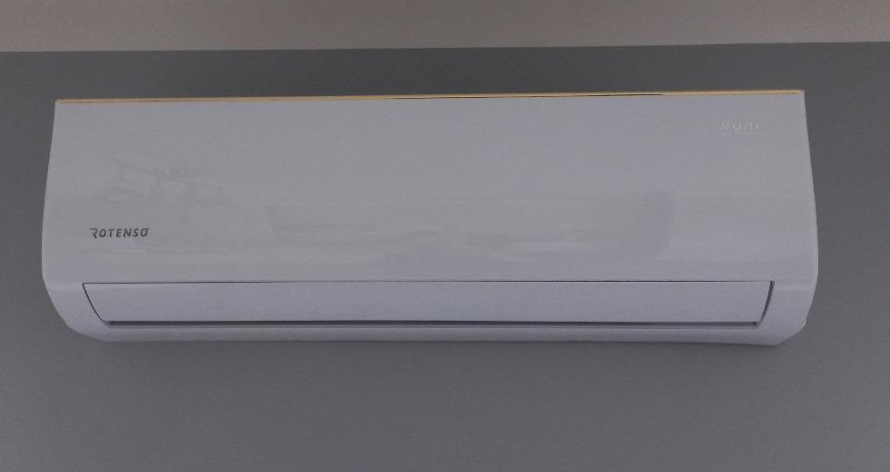 Nowoczesny, biały klimatyzator Rotenso z subtelną złotą linią na górnej krawędzi, zamontowany na szarej ścianie. Minimalistyczny design.