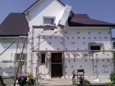 Dom jednorodzinny w trakcie ocieplania styropianem, widoczne rusztowania, częściowo otynkowana elewacja z pozostawionymi punktami mocowania styropianu, brązowy dach, kwiaty w donicach na pierwszym...