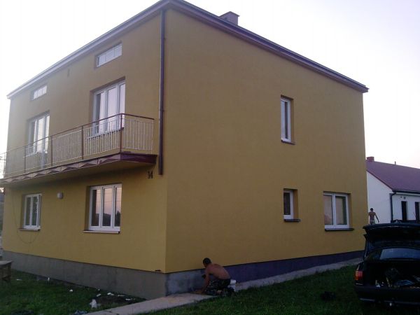 Żółta elewacja dwupiętrowego domu z balkonem, widoczny pracownik przy gruntowaniu fundamentów, otwarty bagażnik samochodu w tle.