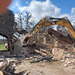 Centracar - Żółta koparka CAT w trakcie rozbiórki budynku z cegły i drewna. Widoczne gruzy, fragmenty dachu i konstrukcji. Błękitne niebo w tle.