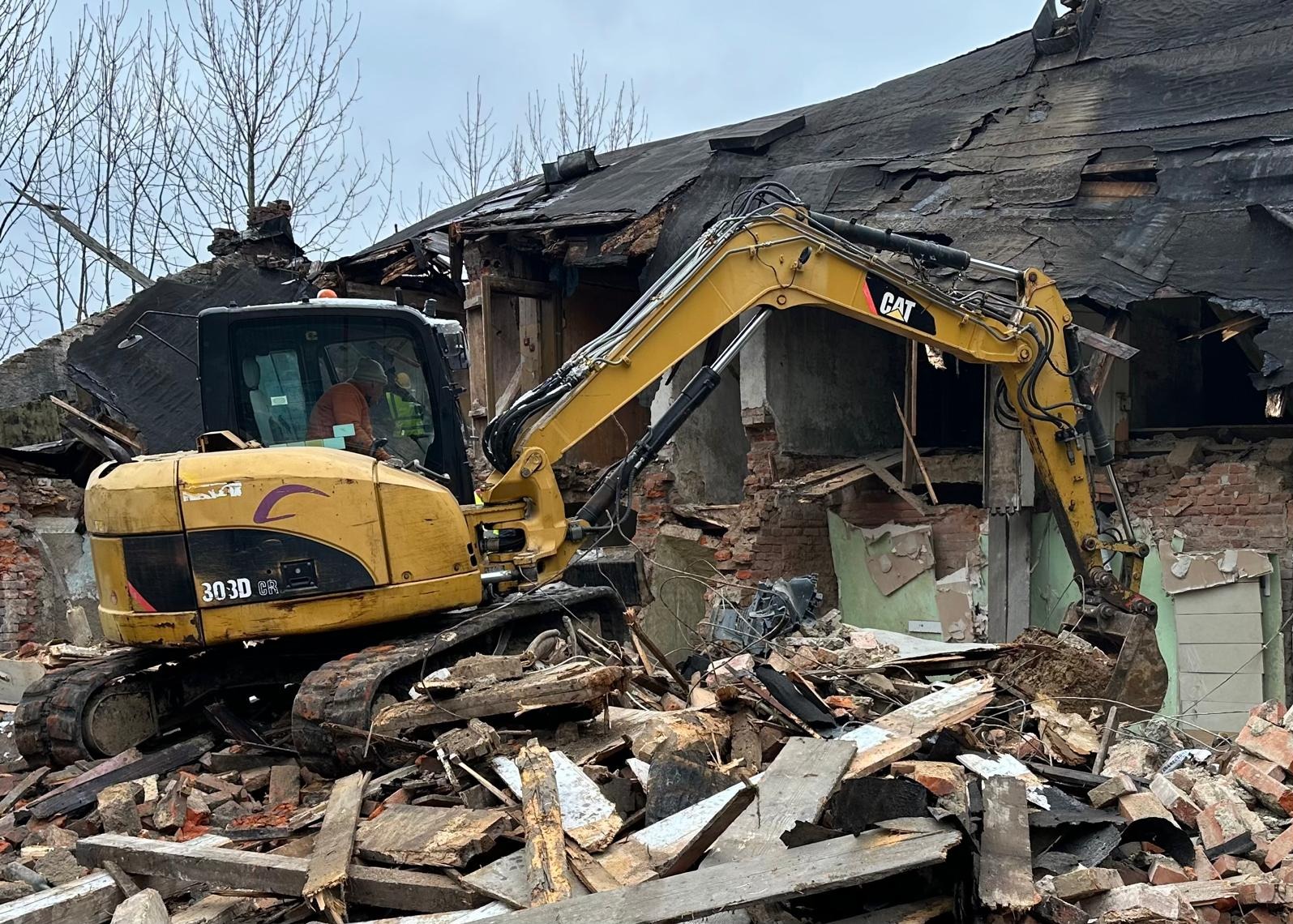 Żółta koparka CAT 303D CR na gruzowisku po rozbiórce budynku. Widoczne fragmenty cegieł, drewna i resztki dachu. Operator w kabinie. Pochmurne niebo w tle.