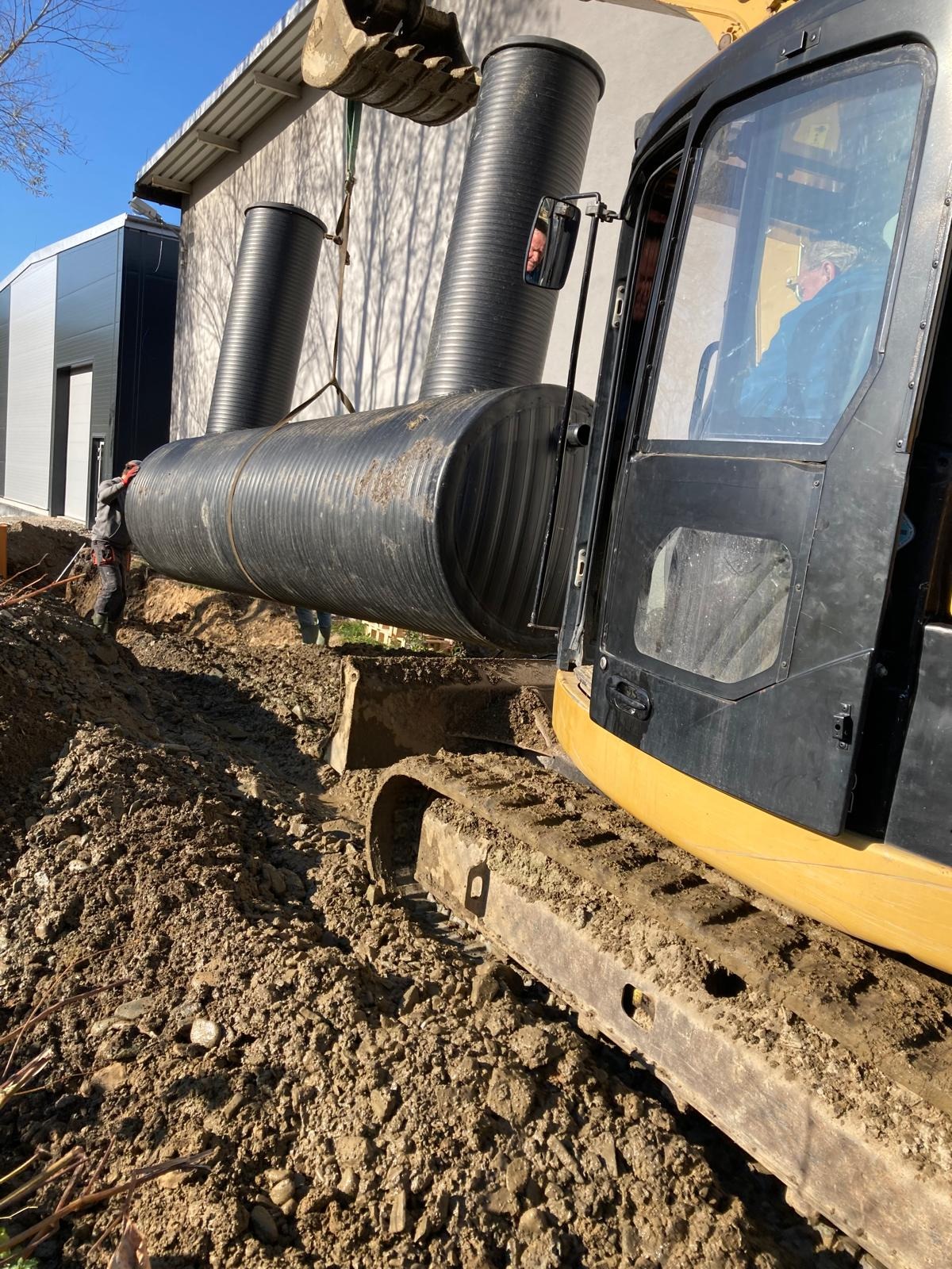 Żółta koparka umieszcza czarny zbiornik w wykopie, pracownik asekuruje. Widoczne elementy budynku i błękitne niebo. Prace ziemne w toku.