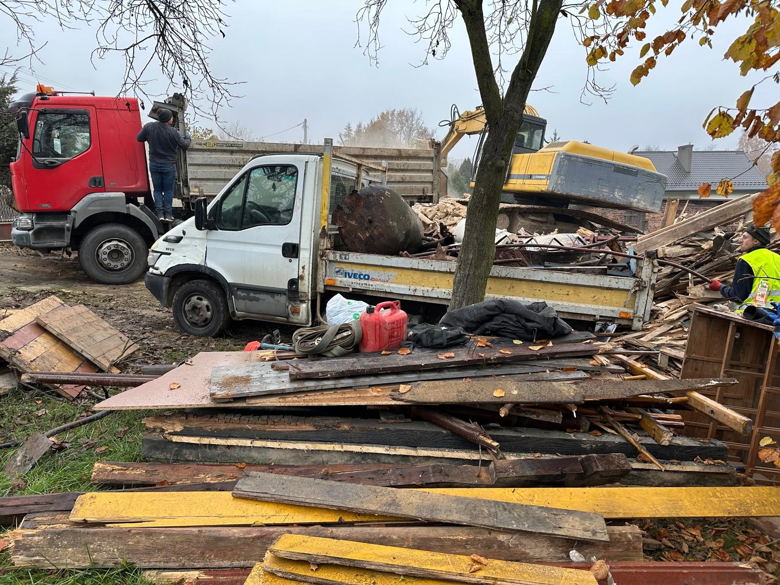 Rozbiórka budynku w Bielsko-Białej: ciężarówki i koparka załadowane gruzem, drewniane elementy w pierwszym planie, pracownik w odblaskowej kamizelce.