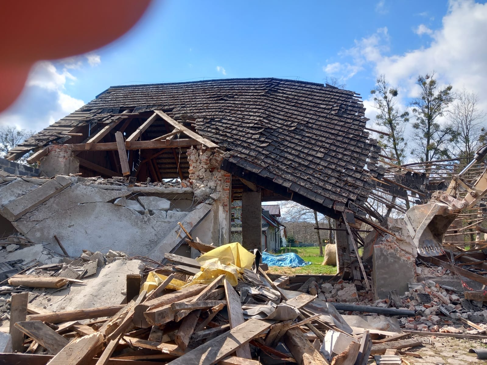 Rozbiórka starego budynku z częściowo zawalonym dachem i stertą gruzu, z widocznym fragmentem koparki i błękitnym niebem w tle.