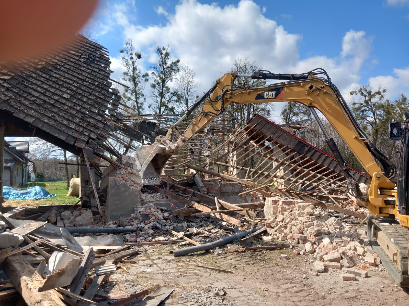 Żółta koparka CAT w trakcie rozbiórki budynku z cegły i drewna. Widoczne gruzy, fragmenty dachu i konstrukcji. Błękitne niebo w tle.