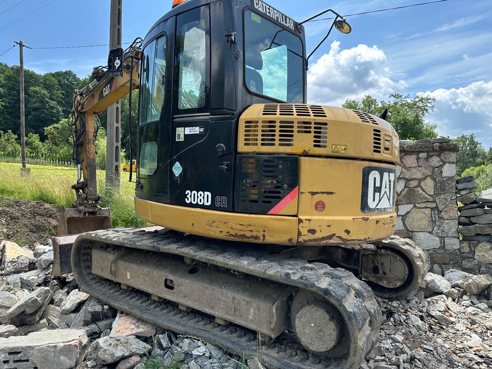 Żółto-czarna koparka CAT 308D CR na gruzowisku, z fragmentem kamiennego muru w tle. Maszyna gotowa do pracy, widoczne ślady użytkowania.