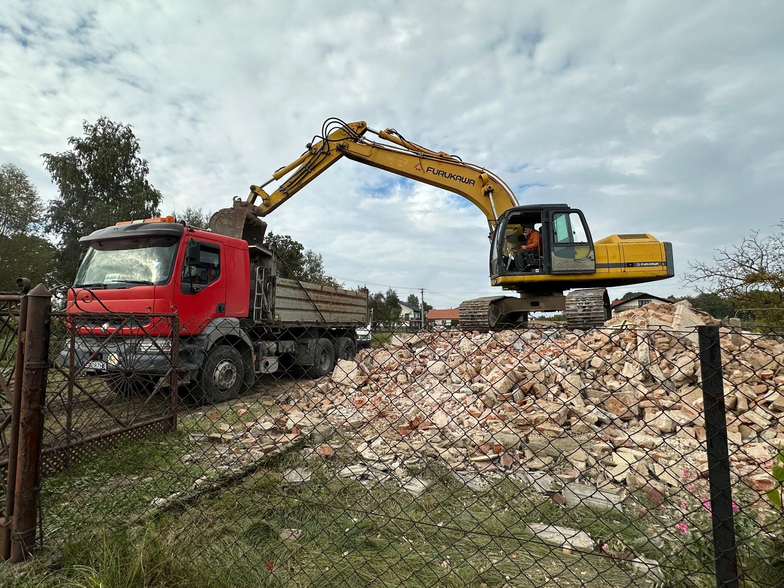 Żółta koparka Furukawa i czerwona ciężarówka na tle gruzu po rozbiórce, widoczne przez metalowe ogrodzenie. Praca w toku, pochmurne niebo.