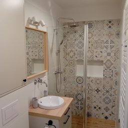 Firma Budowlano-Instalacyjna Bartłomiej Kopacz - Nowoczesna łazienka z kabiną prysznicową, umywalką nablatową i dekoracyjnymi płytkami w odcieniach szarości i beżu. Jasne drewno blatu i podłogi dodaje ciepła.